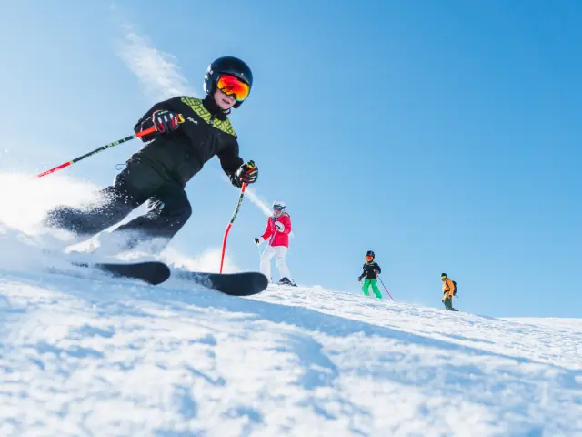 Quatre skieurs, dont des enfants, descendent une piste enneigée sous un ciel parfaitement bleu.