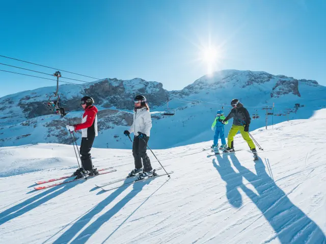Famille skiant ensemble sur une piste ensoleillée du domaine alpin du Dévoluy avec les montagnes enneigées en arrière-plan.