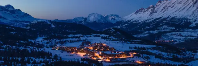 Vue aérienne de la station de Superdévoluy éclairée la nuit, entourée de montagnes enneigées.